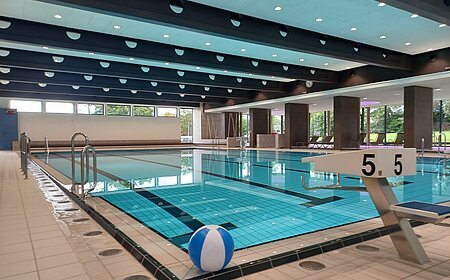 Innenaufnahme des leeren Jurabads mit Becken mit strahlend blauem Wasser. Ein Wasserball liegt dekorativ im Vordergrund.