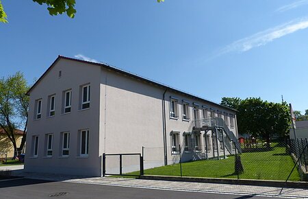 Städtischer Kindergarten in Monheim: ein zweistöckiges Gebäude mit weißer Fassade, Fenstern und Außentreppe, umgeben von einem Zaun und Bäumen. Rechts um das Gebäude ist ein Garten zu sehen. Vorderhalb des Gebäudes verläuft eine Straße.