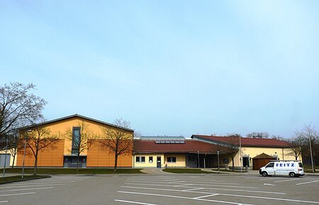 Stadt-/Mehrzweckhalle in Monheim: Ein großes, modernes Gebäude mit gelber Fassade und rotem Dach und mehreren Bäumen. Vorderhalb des Gebäudes ist ein großer Parkplatz, auf dem ein weißes Auto steht.