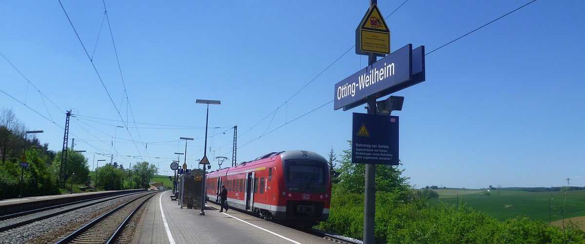 Der Bahnhof Otting-Weilheim mit rotem Zug auf dem Gleis, blauer Himmel, grüne Landschaft im Hintergrund.