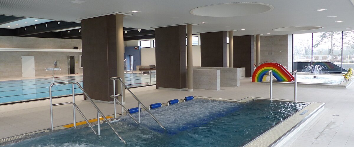 Innenansicht eines Schwimmbads mit mehreren Becken, Rutsche in Regenbogenfarben und Wasserspielen.