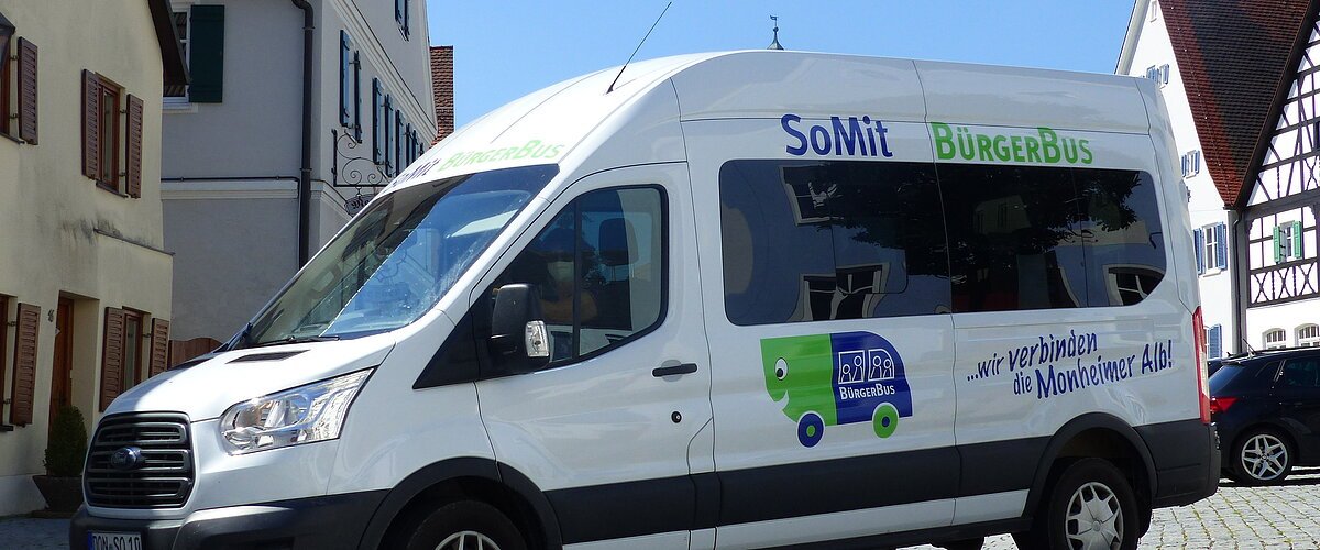 SoMit Bürgerbus Weißer Kleinbus steht auf einem gepflasterten Platz, in der Monheimer Innenstadt . Auf dem Bus ist die Aufschrift "SoMit BürgerBus" und "wir verbinden die Monheimer Alb".
