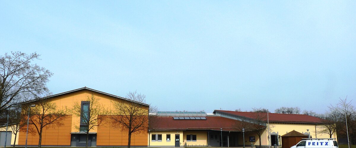 Stadt-/Mehrzweckhalle Monheim Stadt-/Mehrzweckhalle in Monheim: Ein großes, modernes Gebäude mit gelber Fassade und rotem Dach und mehreren Bäumen. Vorderhalb des Gebäudes ist ein großer Parkplatz, auf dem ein weißes Auto steht.