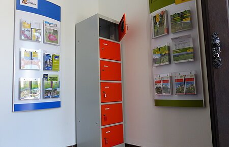 Ein orange-grauer Indoor-Ladeschrank für eBike-Akkus mit insgesamt sechs Fächern und einem offenen Fach steht zwischen zwei Wanddisplays mit Broschüren in dem Vorraum der Touristinformation Monheim.