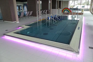 Innenansicht eines Schwimmbades, dem Monheimer Jurabad: ein mit Wasser befülltes Schwimmbecken mit Stufen und Handläufen, umgeben von Fliesenboden. Der Rand des Beckens ist außenrum rosa/lila beleuchtet. Im Hintergrund stehen drei Liegestühle und eine Regenbogenrutsche, sowie ein weiteres Becken.