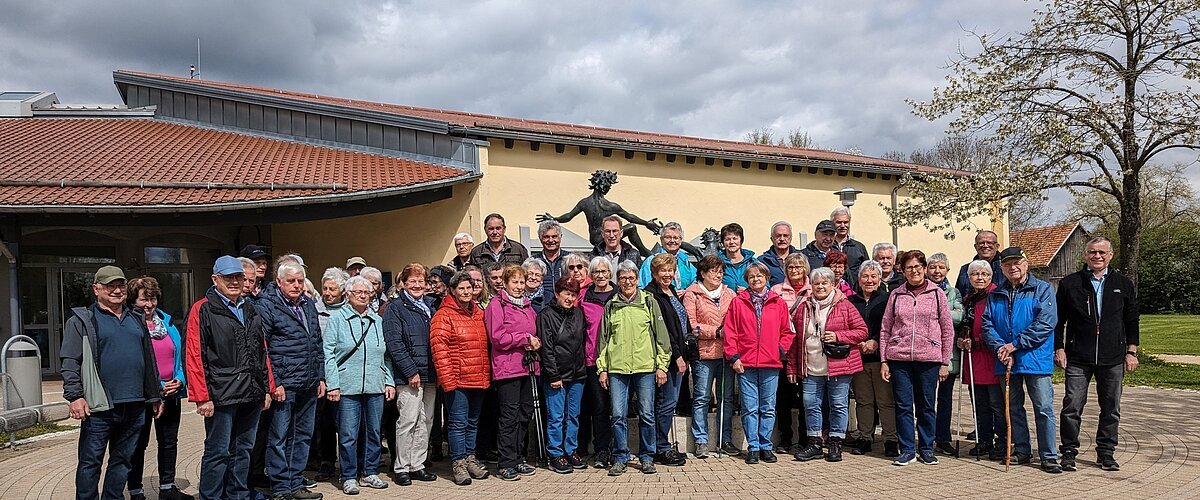 SoMit Wanderung 2024 in Monheim Große Gruppe von Menschen steht vor einem gelben Gebäude mit rotem Dach und einer Statue hinter Ihnen. Im Hintergrund ein Baum auf einer Wiese und bewölkter Himmel.