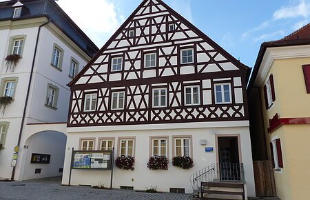 Neue Tourist-Info der Stadt Monheim/Monheimer Alb Tourist-Information der Stadt Monheim/Monheimer Alb: ein Fachwerkhaus mit drei Stockwerken, weißen Wänden und braunen Balken. Davor steht eine Sitzbank und ein Schaukasten auf einem gepflasterten Platz. Links davon ein weißes dreistöckiges Gebäude. Rechts davon ein gelbes zweistöckiges Gebäude mit roten Fensterläden.