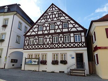Neue Tourist-Info der Stadt Monheim/Monheimer Alb Tourist-Information der Stadt Monheim/Monheimer Alb: ein Fachwerkhaus mit drei Stockwerken, weißen Wänden und braunen Balken. Davor steht eine Sitzbank und ein Schaukasten auf einem gepflasterten Platz. Links davon ein weißes dreistöckiges Gebäude. Rechts davon ein gelbes zweistöckiges Gebäude mit roten Fensterläden.