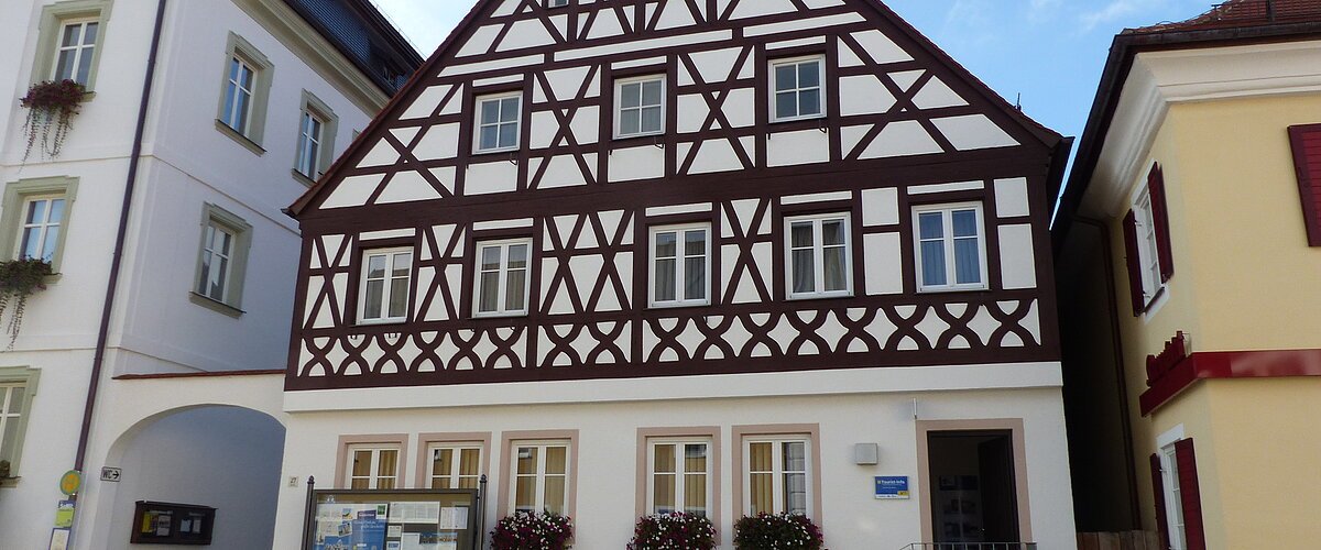 Neue Tourist-Info der Stadt Monheim/Monheimer Alb Tourist-Information der Stadt Monheim/Monheimer Alb: ein Fachwerkhaus mit drei Stockwerken, weißen Wänden und braunen Balken. Davor steht eine Sitzbank und ein Schaukasten auf einem gepflasterten Platz. Links davon ein weißes dreistöckiges Gebäude. Rechts davon ein gelbes zweistöckiges Gebäude mit roten Fensterläden.