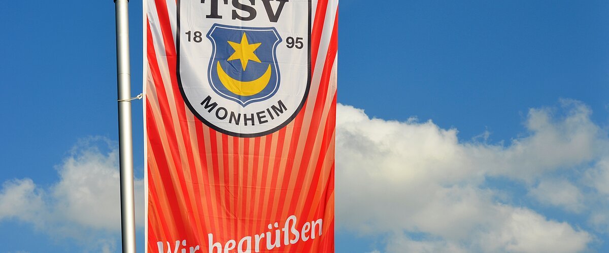 Rote Fahne mit dem Logo des TSV Monheim und dem Text "Wir begrüßen unsere Gäste!" vor blauem Himmel.