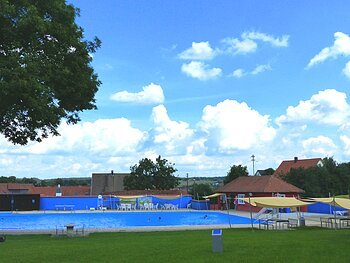 Außenbereich des Monheimer Freibads mit Schwimmbecken, Sonnensegeln, Bäumen und Gebäuden im Hintergrund unter blauem Himmel. Um das Becken grüner Rasen.