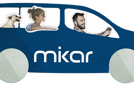 Car-Sharing mikar Blaues Auto mit zwei Personen und einem Hund, auf dem Auto der Schriftzug "mikar" auf der Seite. Fahrer hält das Lenkrad.