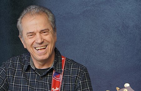 Günther Sigl & Band Pressefoto (c) Dieter Bichl Ein älterer Mann spielt lächelnd Akustikgitarre vor einem blauen Hintergrund.