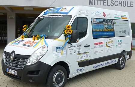 Das neue Carsharing-Modell in Monheim Ein weißer Transporter vom Car-Sharing-Anbieter Mikar mit Werbeaufdrucken und Sonnenblumen steht vor einem Gebäude mit der Aufschrift "Grundschule Mittelschule" in Monheim.