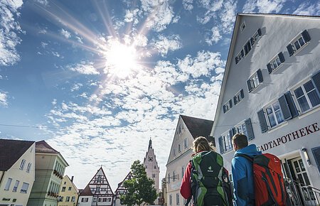 Wanderer in Monheim Zwei Personen mit Rucksäcken gehen durch eine Stadt mit Fachwerkhäusern unter einem sonnigen Himmel. Auf dem Haus im Vordergrund steht die Aufschrift "Stadtapotheke".