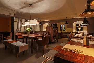 Ein Restaurant mit Holztischen, Stühlen und gedämpfter Beleuchtung. Auf den Tischen Besteck und Gewürzbehälter und auf einem Tisch stehen Blumen in der Vase.