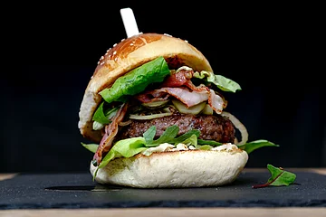 Burger- Brötchen gefüllt mit Salat, Speck, Gurken und Zwiebeln, auf schwarzem Hintergrund und einer schwarzen Servierplatte.