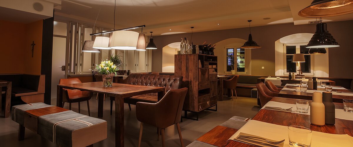 Ein Restaurant mit Holztischen, Stühlen und gedämpfter Beleuchtung. Auf den Tischen Besteck und Gewürzbehälter und auf einem Tisch stehen Blumen in der Vase.