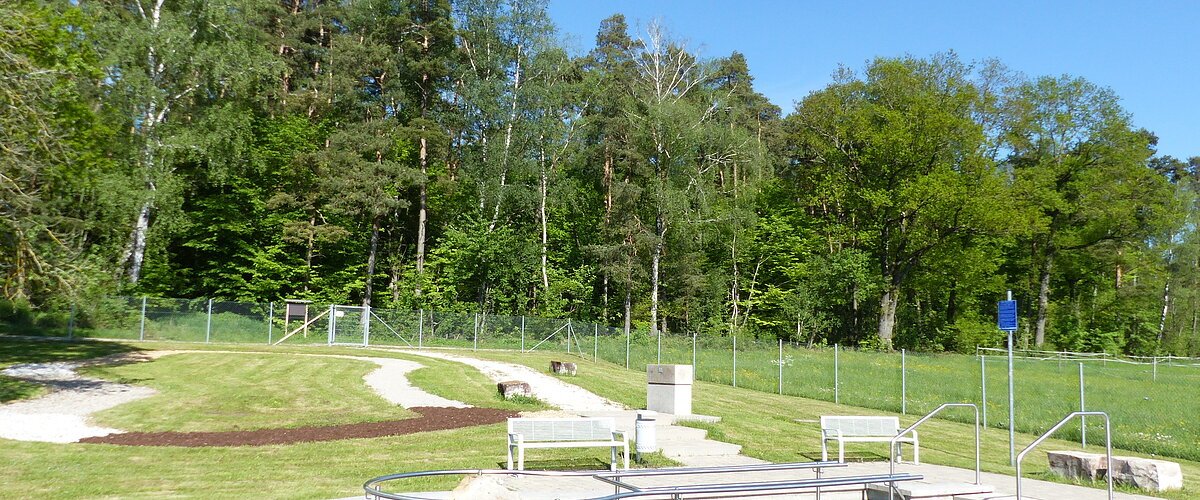 Rundes Kneippbecken in Monheim mit einem Geländer und einem Barfußpfad auf einer Wiese, umgeben von einer großen Grünfläche und Bäumen und Sitzmöglichkeiten. Ein Zaun im Hintergrund.