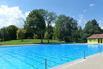 Freibad mit blauer Wasseroberfläche, im Hintergrund Bäume und ein gelbes Gebäude.