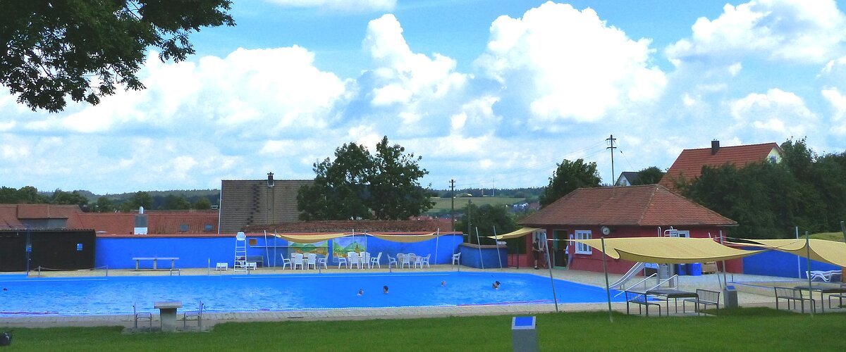 Freibad mit blauem Wasser, umgeben von Rasen, Bäumen und Gebäuden unter einem bewölkten Himmel. Vorderhalb vom Becken sind Sitzmöglichkeiten.