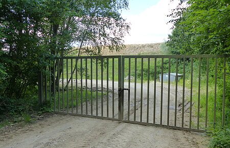 Erdaushubdeponie Monheim Verschlossenes Metalltor auf einem unbefestigten Weg, umgeben von Bäumen und Gras. Im Hintergrund ist ein Müllcontainer und ein Hügel sichtbar.