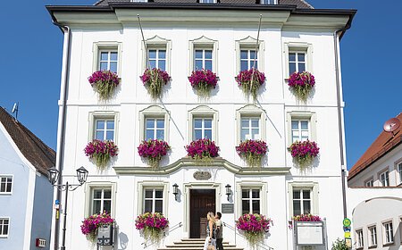 Das Rathaus in Monheim: Ein weißes dreistöckiges Gebäude mit mehreren Fenstern, an denen Blumenkästen mit pinken Blumen hängen. Zwei Personen gehen davor vorbei. Links und rechts davon jeweils ein weiteres Haus.