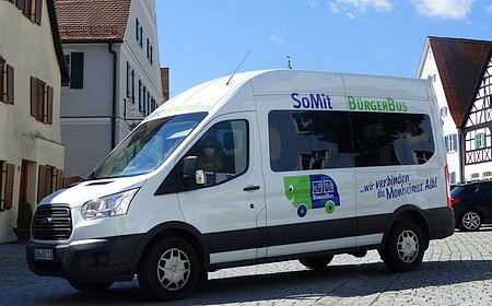 Weißer Kleinbus steht auf einem gepflasterten Platz, in der Monheimer Innenstadt . Auf dem Bus ist die Aufschrift "SoMit BürgerBus" und "wir verbinden die Monheimer Alb".
