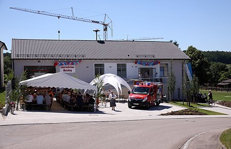 Neues Feuerwehrhaus Warching, Schloßberg 4 Feuerwehrhaus mit einem weißen Festzelt, einem weißen Pavillon und einem Feuerwehrfahrzeug davor. Menschen sitzen unter dem Zelt, ein Kran steht hinter dem Feuerwehrhaus, im Vordergrund eine gepflasterte Straße. Feuerwehrhaus ist umgeben von Feldern und einem Wald.