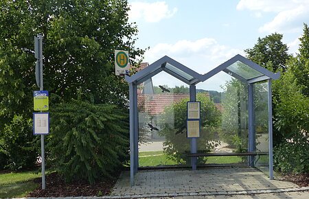 Bushaltestelle Weilheim Bushaltestelle mit Glasüberdachung, umgeben von Bäumen und Sträuchern, mit Fahrplan und Haltestellenschild.