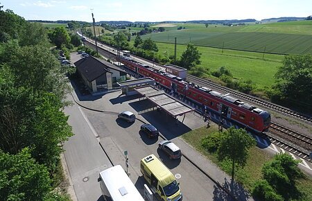 Bahnhof Otting-Weilheim Luftaufnahme eines Bahnhofs mit rotem Zug, umgeben von Feldern und Bäumen. Stehende Autos und Busse sind zu sehen.