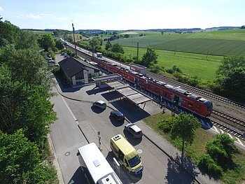 Bahnhof Otting-Weilheim Luftaufnahme eines Bahnhofs mit rotem Zug, umgeben von Feldern und Bäumen. Stehende Autos und Busse sind zu sehen.
