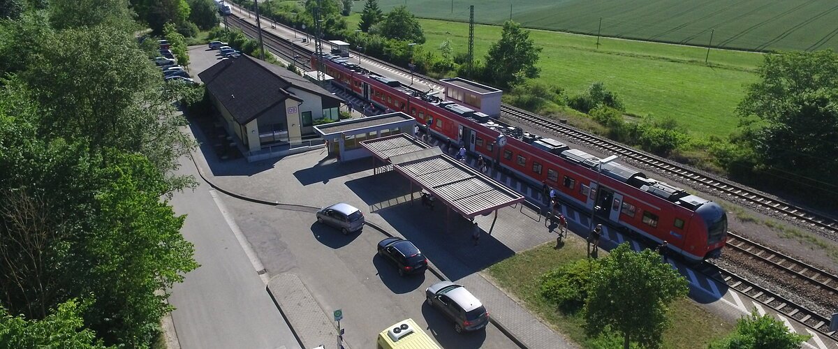 Luftaufnahme eines Bahnhofs mit rotem Zug, umgeben von Feldern und Bäumen. Stehende Autos und Busse sind zu sehen.