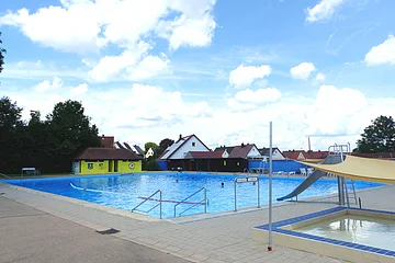 Ein Freibad mit einem Schwimmbecken, einer Rutsche und einem Kinderbecken. Im Hintergrund steht ein grünes Gebäude mit rotem Dach. Dahinter sind Wohnhäuser und Bäume und blauer Himmel zu sehen.