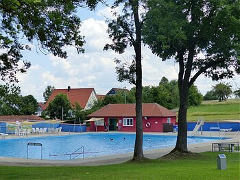 Ein Freibad mit Schwimmbecken, einer Rutsche und Sitzmöglichkeiten, umgeben von Bäumen und Wiesen. Im Hintergrund steht ein rotes Gebäude mit Fenstern, ein Kiosk.
