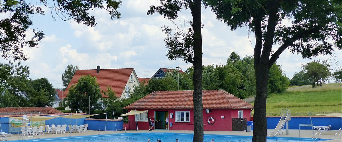 Ein Freibad mit Schwimmbecken, einer Rutsche und Sitzmöglichkeiten, umgeben von Bäumen und Wiesen. Im Hintergrund steht ein rotes Gebäude mit Fenstern, ein Kiosk.