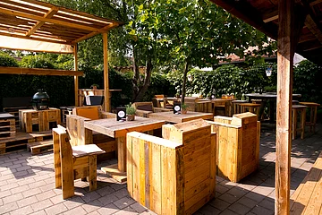Holztische- und Stühle auf einer Terrasse mit Pergola und Pflanzen im Hintergrund, auf einer gepflasterten Fläche.