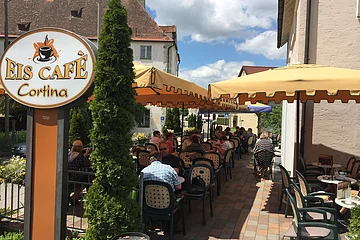 Außenbereich eines Eiscafés mit gelben Sonnenschirmen, Tischen und Stühlen, Gäste sitzen und essen.