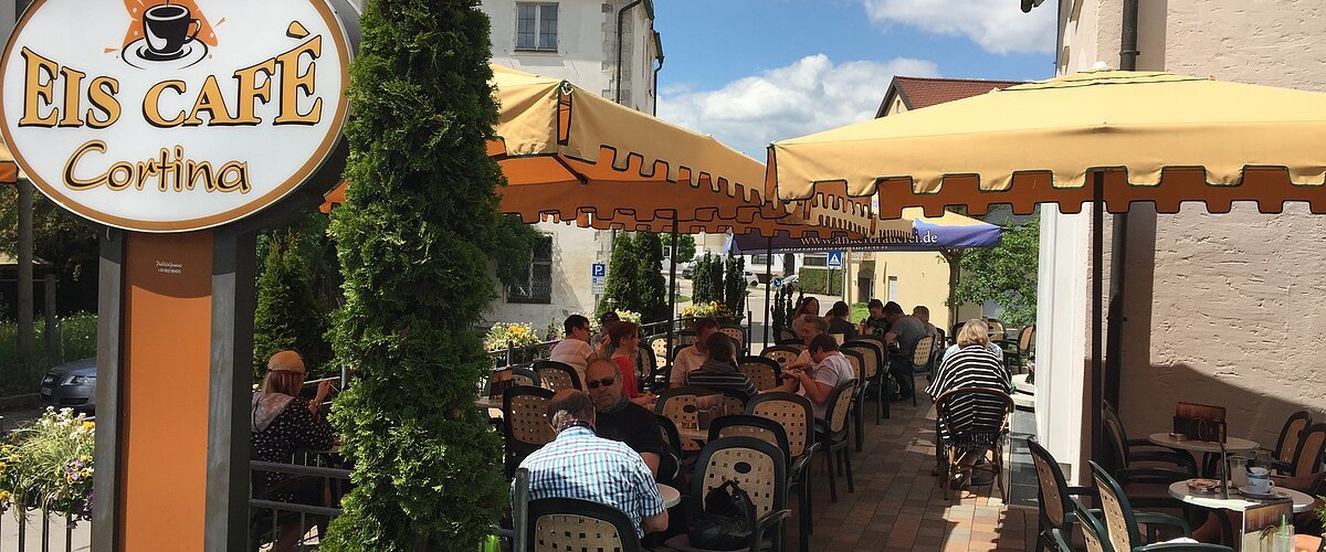 Außenbereich eines Eiscafés mit gelben Sonnenschirmen, Tischen und Stühlen, Gäste sitzen und essen.
