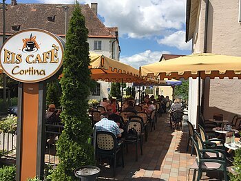 Außenbereich eines Eiscafés mit gelben Sonnenschirmen, Tischen und Stühlen, Gäste sitzen und essen.