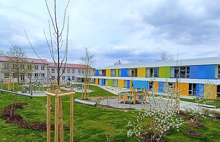 Städtische Kindertagesstätte Monheim Zwei Gebäude des städtischen Kindergartens Monheim, eins davon mit einer bunten Fassade, davor ein Garten mit Spielgeräten Bäumen und Rasenfläche, bewölkter Himmel.