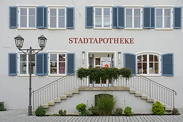 Gebäude mit blauen Fensterläden, rotem Schriftzug "STADTAPOTHEKE", einer Treppe und einer Laterne mit einem bepflanzten Beet davor.