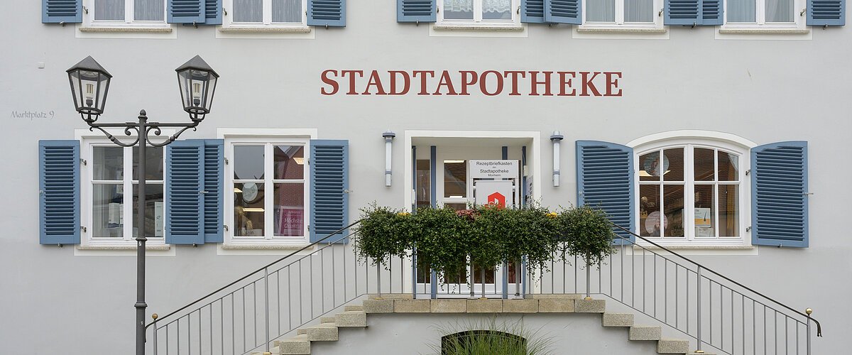 Gebäude mit blauen Fensterläden, rotem Schriftzug "STADTAPOTHEKE", einer Treppe und einer Laterne mit einem bepflanzten Beet davor.