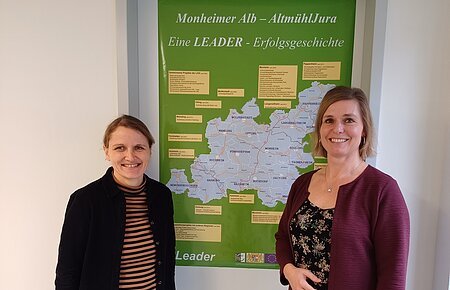 LAG Geschäftsstelle Monheim Zwei lächelnde Personen stehen vor einem grünen Plakat mit einer Karte der Region Monheimer Alb-AltmühlJura. Umgeben von einer weißen Wand.