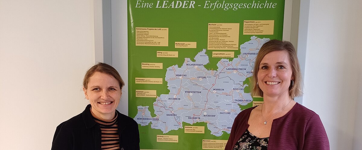 Zwei lächelnde Personen stehen vor einem grünen Plakat mit einer Karte der Region Monheimer Alb-AltmühlJura. Umgeben von einer weißen Wand.