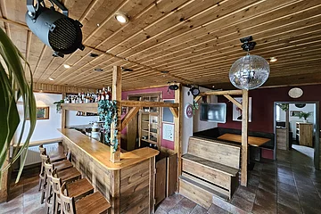 Ein rustikaler Barbereich mit Holzmöbeln, einer Theke, Barhockern, einem schwarzen Scheinwerfer und einer Discokugel an der Decke.