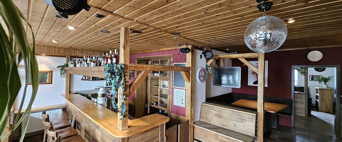 Ein rustikaler Barbereich mit Holzmöbeln, einer Theke, Barhockern, einem schwarzen Scheinwerfer und einer Discokugel an der Decke.