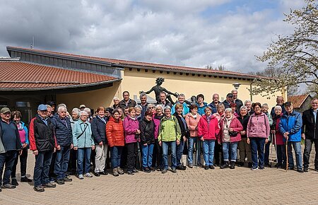 SoMit Wanderung 2024 in Monheim Große Gruppe von Menschen steht vor einem gelben Gebäude mit rotem Dach und einer Statue hinter Ihnen. Im Hintergrund ein Baum auf einer Wiese und bewölkter Himmel.