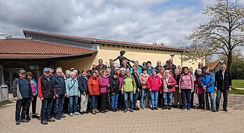 Große Gruppe von Menschen steht vor einem gelben Gebäude mit rotem Dach und einer Statue hinter Ihnen. Im Hintergrund ein Baum auf einer Wiese und bewölkter Himmel.