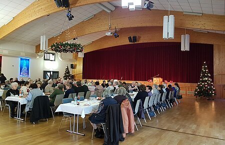 Seniorenweihnachtsfeier Stadt Monheim & Bayer. Rotes Kreuz 2017 Menschen sitzen in mehreren Reihen an weißgedeckten Tischen in einem Saal mit einer Holzbalkendecke und Weihnachtsdekoration. Eine Bühne mit einem roten Vorhang ist im Hintergrund.